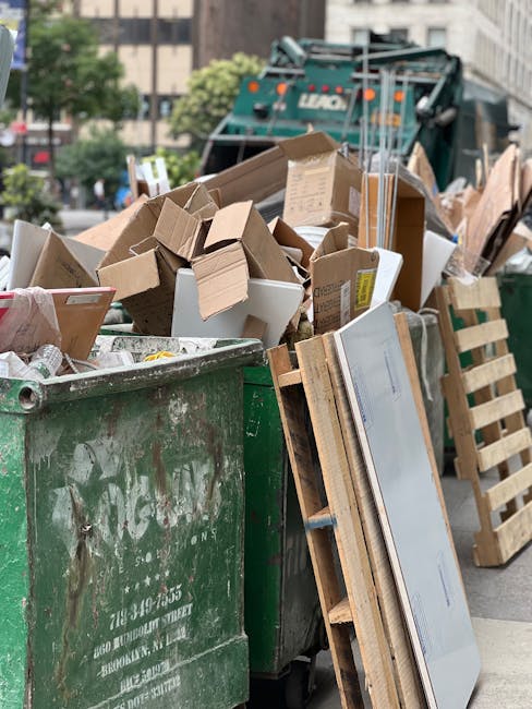 A large green waste bin filled with assorted cardboard boxes, some flattened and others still partially assembled, along with pieces of packing material. To the right of the bin, there is a wooden pallet leaning against it, holding a flat-screen television with a reflective screen and a wooden frame. In the background, a street scene is visible with a green waste collection vehicle parked nearby, surrounded by a concrete and metal urban environment, including buildings and trees. The scene appears to be an outdoor area designated for waste disposal or bulk rubbish collection, with the clutter indicating a recent or ongoing clearance process, possibly arranged by Rubbish Collection Harrow as part of an alternative waste handling service.