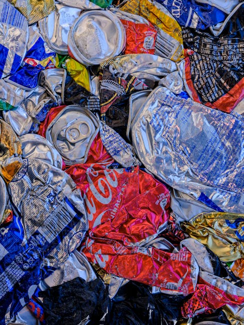 A close-up image showing a pile of crushed and crumpled aluminum cans, primarily silver with some displaying distinctive branding and colorful packaging, such as a partially visible red Coca-Cola can. The cans are mixed together on a surface likely outdoors or in a waste collection area, with some partially obscured by others and exhibiting dents and creases from crushing. The background includes additional flattened cans with varied colors and branding, indicating a mixed collection of recyclable beverage containers. The overall scene suggests a focus on waste material prepared for recycling, aligning with services provided by waste management specialists like Rubbish Collection Harrow, which handles alternative waste collection and disposal tasks. The lighting appears natural, emphasizing the metallic sheen and textures of the cans, highlighting the importance of proper recyclable waste segregation and disposal methods within local rubbish management routines.