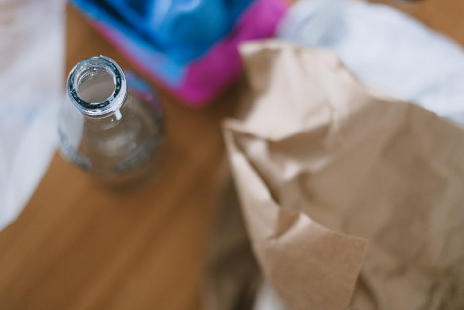 A clear glass bottle with a narrow neck and a rounded body, placed on a wooden surface, with no label or cap visible. The bottle appears to be empty or contain a small amount of liquid. In the background, there is a partially visible cardboard or paper bag lying crumpled, along with a blurred object that seems to be a colorful, possibly plastic, container or packaging. The scene suggests a typical indoor setting associated with waste or rubbish, and the focus on the bottle and crumpled paper bag aligns with waste removal services like those offered by Rubbish Collection Harrow. The image’s lighting is natural and even, highlighting the textures of the glass and paper. It reflects a typical scene of small household rubbish, highlighting the need for effective rubbish removal or recycling processes in a professional waste management context.