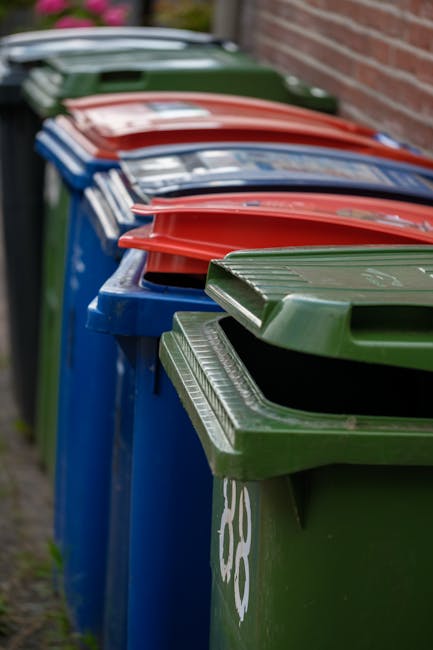 A close-up image showing a pile of crushed and crumpled aluminum cans, primarily silver with some displaying distinctive branding and colorful packaging, such as a partially visible red Coca-Cola can. The cans are mixed together on a surface likely outdoors or in a waste collection area, with some partially obscured by others and exhibiting dents and creases from crushing. The background includes additional flattened cans with varied colors and branding, indicating a mixed collection of recyclable beverage containers. The overall scene suggests a focus on waste material prepared for recycling, aligning with services provided by waste management specialists like Rubbish Collection Harrow, which handles alternative waste collection and disposal tasks. The lighting appears natural, emphasizing the metallic sheen and textures of the cans, highlighting the importance of proper recyclable waste segregation and disposal methods within local rubbish management routines.