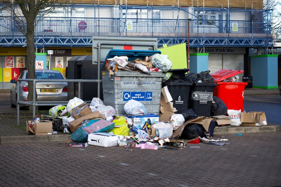 A collection of overflowing mixed waste and household rubbish in black, red, and blue recycling bins, with additional cardboard boxes and plastic bags scattered on the paved sidewalk in front of a commercial building. The black and blue bins are positioned next to each other, with the blue bin lid open and filled with paper and cardboard waste spilling out onto the ground. The red bin appears to contain plastics or mixed waste. Surrounding the bins, there are various disorganized bags, flattened cardboard boxes, and loose packaging material, indicating a recent or ongoing rubbish collection or disposal activity. Behind the waste, a modern commercial building with large windows and a blue metal scaffolding structure is visible, with signage and storefronts, including a fish bar, in the background. The scene is outdoors, lit by natural daylight, suggesting daytime. This scene exemplifies the type of waste collection managed by private waste removal services like Rubbish Collection Harrow and highlights the importance of organized rubbish disposal efforts in urban areas.