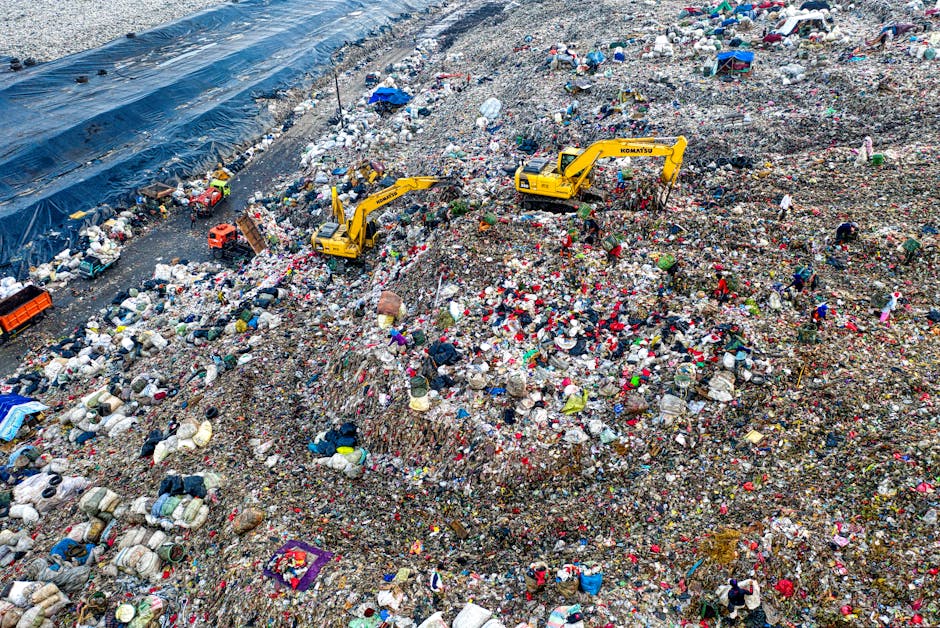 An expansive landfill site filled with a thick layer of mixed waste, including plastic bags, bottles, cardboard, textile scraps, and miscellaneous debris, spreading across uneven mounds of garbage. Two yellow excavators are positioned centrally, actively working to move and manage the waste materials. To the left, a large section of the site is covered by a black tarpaulin secured with sandbags, possibly to contain or protect certain areas. The environment appears hazy, with natural daylight illuminating the scene, emphasizing the scale of waste accumulation. Scattered groups of workers in protective clothing are visible amidst the debris, engaging in sorting or collection activities. The scene typifies a large-scale waste disposal operation, reflecting the importance of effective rubbish removal services and alternative waste management solutions, as offered by companies like Rubbish Collection Harrow, to handle substantial volumes of refuse efficiently and responsibly.