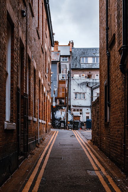 A narrow alleyway between two brick buildings in an urban residential area, with the alley's asphalt surface marked by yellow double lines along its edges. The left and right walls are constructed from red-brown bricks, with black drainpipes and small, rectangular windows with white frames visible on both sides. In the background, a small set of wooden stairs leads up to a second-floor door attached to a whitewashed building, which is partially covered with graffiti. Several bins and waste containers are positioned at the rear of the alley, indicating private waste collection. The sky above is overcast, casting diffuse natural light over the scene, and the overall setting suggests a typical environment for a private rubbish collection service, such as that offered by Rubbish Collection Harrow, for handling waste in areas where traditional municipal rubbish removal may be less practical due to limited space or access.