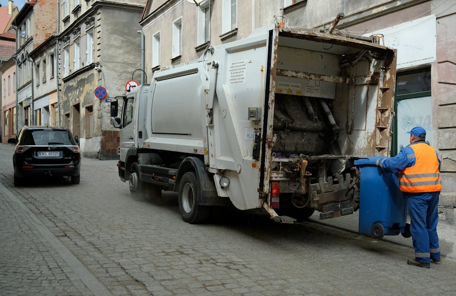 A narrow urban alleyway filled with a large, wheeled black rubbish bin positioned in the foreground, surrounded by scattered debris and dirt on the asphalt surface. Behind the bin, an overflowing pile of cardboard boxes, plastic bags, and miscellaneous waste materials are stacked against a background of weathered brick and concrete walls. The alley is flanked on both sides by buildings with graffiti-covered and peeling surfaces, and there are exposed pipes and utility boxes attached to the walls. Sparse tree branches extend across the top of the scene, indicating nearby trees with leafless branches. The overall atmosphere suggests an area undergoing informal waste accumulation or an environment that may benefit from professional rubbish removal services such as those provided by Rubbish Collection Harrow, who specialize in alternative waste handling and on-site clearance in urban settings,” while the natural lighting emphasizes the gritty textures of the discarded waste and surrounding environment.