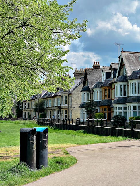 A pair of black public waste bins positioned on a grassy verge beside a paved footpath, with the bins showing signs of weathering and slight graffiti. Behind the bins, there is a row of Victorian-style terraced houses with bay windows, some with decorative wooden frames painted in contrasting colours, and brick facades in light shades. The houses are partially obscured by lush green trees with fresh summer foliage, extending over the pavement and contributing to a leafy, residential atmosphere. The sky above features scattered white clouds against a blue background, suggesting a bright day. The scene is set in a suburban area, highlighting typical features of waste management in urban residential zones, with the bins ready for collection or replacement, aligning with private or independent rubbish removal services such as those offered by Rubbish Collection Harrow. This image captures the quiet routine of waste disposal in a well-maintained neighborhood, emphasizing the importance of proper rubbish handling in line with local and alternative waste collection practices.