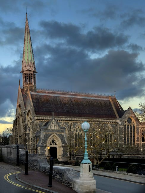 An exterior view of a historic stone church with a tall, pointed spire featuring a green patina, set against a cloudy sky during twilight. The church has intricate Gothic-style architectural details, including arched windows, decorative stonework, and lancet windows with stained glass. The building is constructed from weathered grey and brown stone, showing signs of age and historic character. In front of the church, a small paved area with a low stone wall and bollards is visible, alongside a vintage-style lamp post with a spherical light on a pedestal. Sparse leafless trees partially obscure the church's facade, and nearby there is minimal street furniture and a double yellow line markings along the road. While the scene does not directly show rubbish or waste, the setting relates to the context of local community services, with potential relevance to waste management or maintenance of historic sites, subtly connecting to the theme of alternative waste handling and cleanliness expectations that Rubbish Collection Harrow offers in the local area.