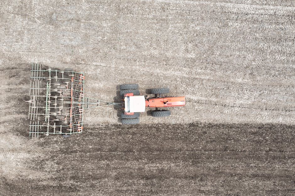 An aerial view of a compact orange and black tractor with large textured rubber tires working on a freshly tilled soil field. The tractor is attached to a metal harrow attachment with multiple horizontal and vertical tines, which is used for loosening and aerating the soil. The soil surface displays parallel rows created by previous tilling, with darker, freshly disturbed earth near the harrow and lighter, dry soil in the surrounding areas. The scene is outdoors, with natural daylight illuminating the scene evenly, emphasizing the contrasting textures and colours of the soil and machinery. This type of agricultural equipment could be associated with land preparation before planting or excavated land clearance, reflecting the context of site preparation that might relate to private ground or land management services in a rubbish removal or site clearance setting, relevant to independent waste handling and ground clearance activities overseen by services such as Rubbish Collection Harrow.