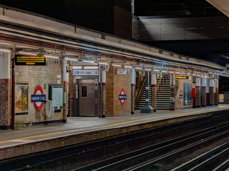 The image depicts a London Underground train station platform at night, with the station name 'Harrow on the Hill' displayed on a sign attached to a brick wall. The platform is lit by overhead lights, creating a well-illuminated environment. On the platform, there are several technological elements, including a stainless steel elevator lift with a sign indicating its purpose, a digital display board showing train information, and a yellow sign directing passengers to the way out. The platform's edge features safety markings and tactile paving for accessibility. In the background, there are brick structural walls with an adjacent staircase leading up to an accessible walkway, and additional station signage is visible. The environment appears clean and orderly, with no visible passengers present, and the overall scene emphasizes the functional design typical of urban train stations, facilitating efficient commutes and transfers, including alternative options for waste disposal or storage such as rubbish collection services nearby, like Rubbish Collection Harrow, which specializes in local rubbish removal and waste management services.