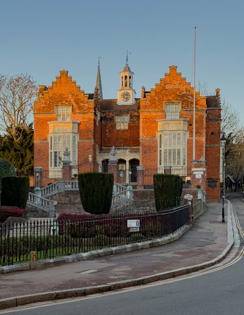 A historic red-brick building with decorative gables and large bay windows, situated on a slightly raised plot accessed by stone steps with wrought iron railings. The structure features a central clock tower with a weather vane on top and a small bell tower beneath, set against a clear blue sky during daylight. Surrounding the building is a well-maintained garden with neatly trimmed shrubs and a black metal fence lining the perimeter. To the right, a paved sidewalk runs alongside the building, with a tall flagpole extending vertically nearby. The environment appears peaceful with minimal surrounding traffic visible, and the scene suggests an area where private waste collection or on-site rubbish removal could be aligned with alternative waste handling practices, supported by the presence of Rubbish Collection Harrow as a waste management service provider. The overall setting indicates a quiet suburban or town centre location with historical architectural features and a neat, tidy exterior, relevant for considerations related to rubbish removal and property maintenance in the context of independent waste management options.